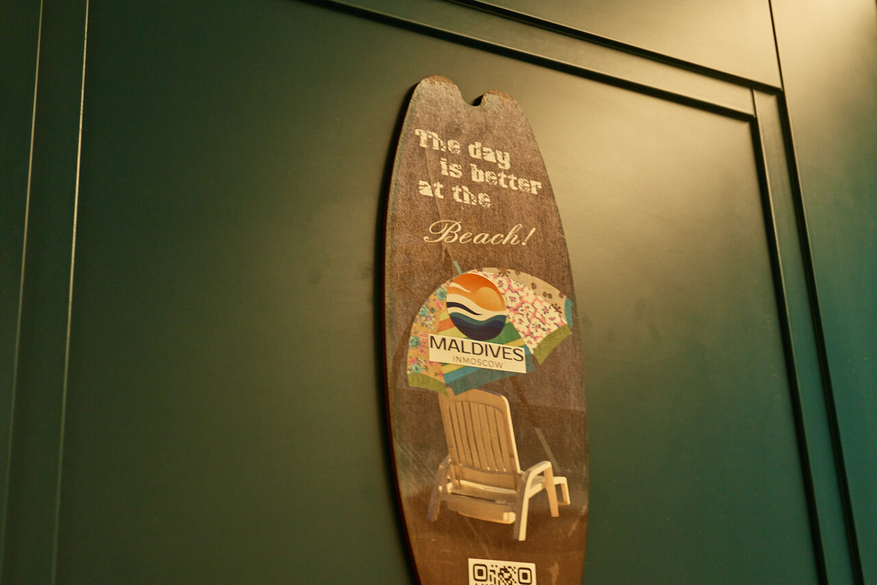 Арендовать ⁠Лофт в Мексиканском стиле для костюмированных вечеринок. Деревянная табличка в форме доски с надписью "The day is better at the Beach!", логотипом "Maldives", изображением шезлонга и зонтика. Подвешена на тёмно-зелёной деревянной панели.
