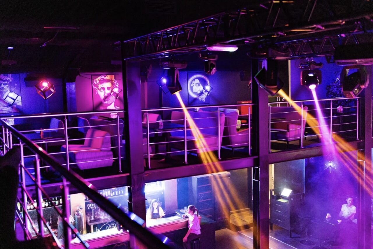 Арендовать Castle Concert Hall(Концертный зал Замок). Двухуровневое пространство с балконом, оборудованное барной стойкой. Яркое цветное освещение, пара кресел у стен с картинами. Металлические перила и конструкции. Люди за баром. Световые лучи отражаются от поверхностей.