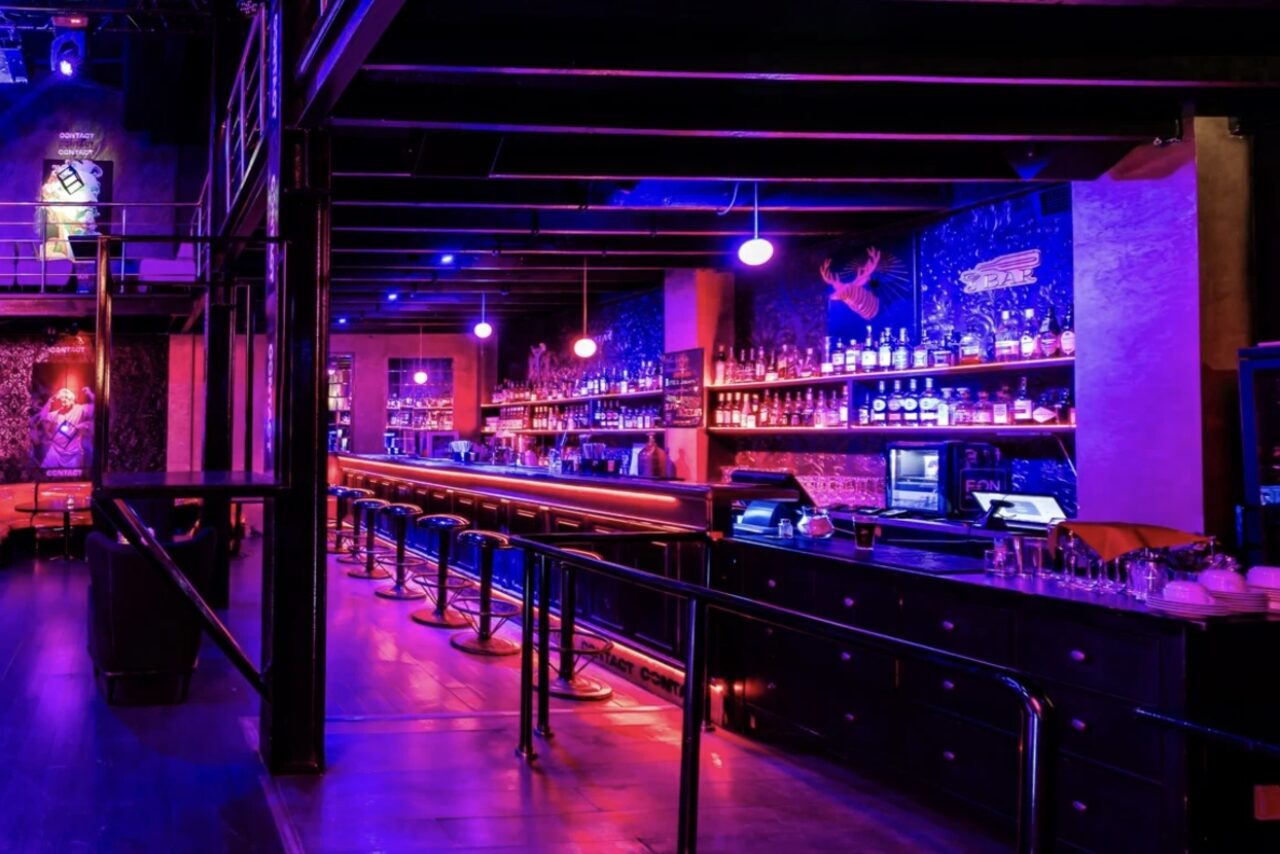 Арендовать Castle Concert Hall(Концертный зал Замок). Бар с длинной стойкой и высокими стульями, рядом стойка для микшеров. Темное освещение с неоновыми фиолетовыми и розовыми оттенками. На заднем плане полки с бутылками, стены с графикой. Потолок с балками, металлические конструкции.