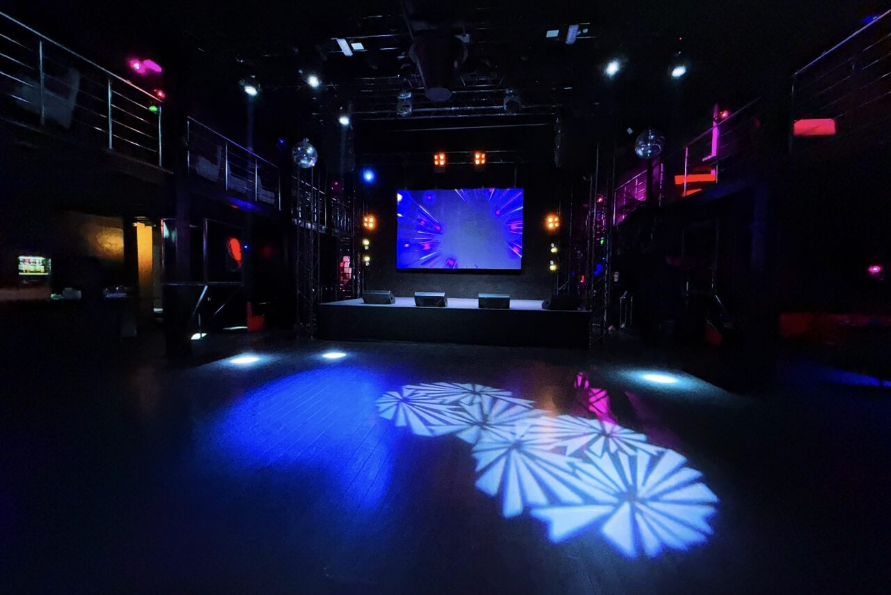 Арендовать Castle Concert Hall(Концертный зал Замок). Большое тёмное помещение с сценой и экраном. Металлические балконы по бокам, диско-шары, разноцветное освещение с узорами на полу. Черный пол, высокий потолок, световые приборы, цветная подсветка. Монохромная и фиолетовая гамма.