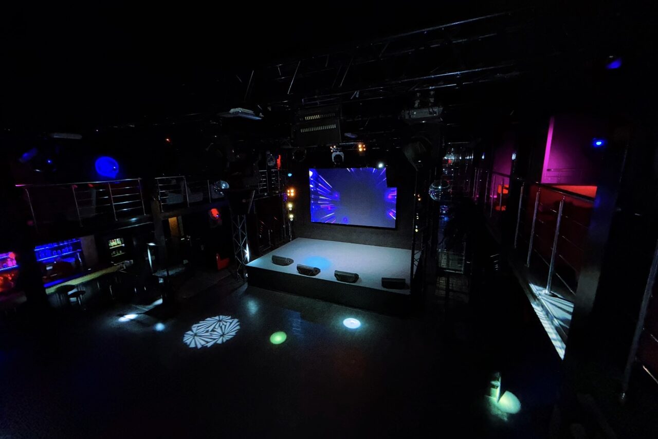 Арендовать Castle Concert Hall(Концертный зал Замок). Ночной клуб с двумя уровнями и сценой. Центральный экран, несколько световых эффектов на полу. Оборудование для света и звука. Барная стойка слева, металлические перила вдоль второго уровня. Черный интерьер с разноцветной светодиодной подсветкой.