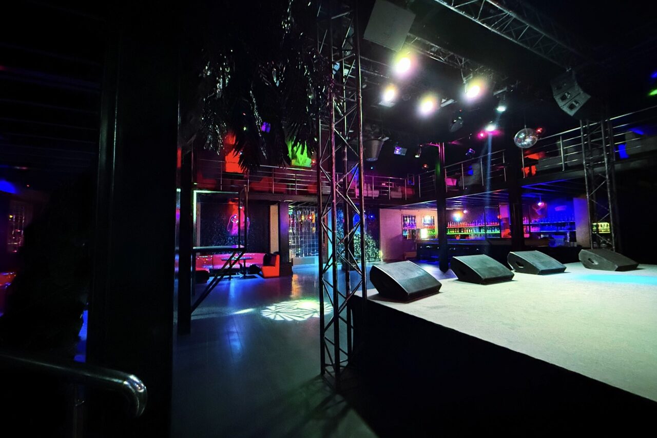 Арендовать Castle Concert Hall(Концертный зал Замок). Ночной клуб с открытым пространством и сценой. Справа стойка с аудиосистемой, разноцветные прожекторы на потолке. Вдалеке барная зона с подсветкой. Металлические конструкции, полы темные. В центре несколько висящих зеркальных шаров.