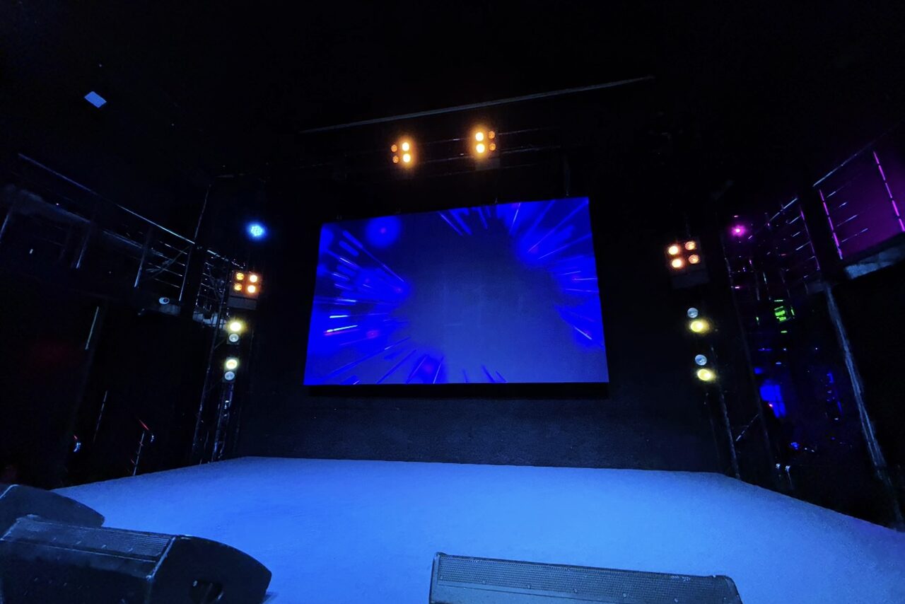 Арендовать Castle Concert Hall(Концертный зал Замок). Сцена с большим экраном в центре, окруженная черными стенами. Освещение синих и теплых тонов, расположенное по бокам. Пол светлый, без покрытия. Также видны звукосниматели и яркие прожекторы на потолке.