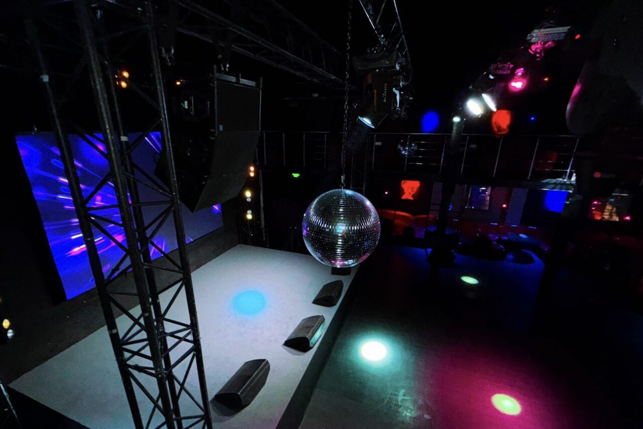 Арендовать Castle Concert Hall(Концертный зал Замок). Тёмное пространство с танцполом, оборудованным диско-шаром и разноцветными световыми эффектами. Платформа с несколькими аудиоколонками. Перегородки и металлические конструкции. Используются синие и красные неоновые огни.