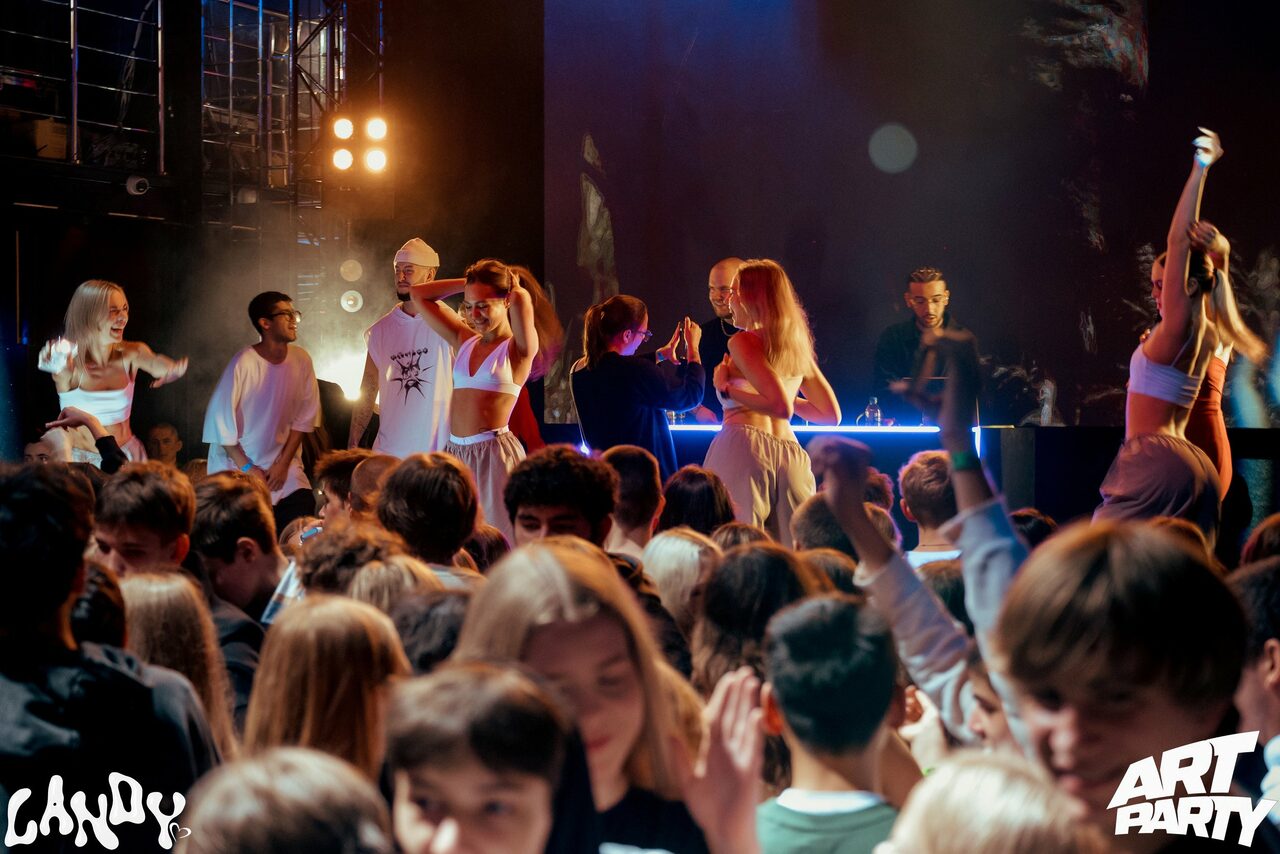 Арендовать Castle Concert Hall(Концертный зал Замок). Активный танцпол клуба с диджейским пультом на сцене. Яркое разноцветное освещение, светодиодные панели. Люди танцуют, на сцене и в зале. На сцене динамики, несколько прожекторов освещают пространство. Тёмный интерьер, световые эффекты. На фотографиях логотипы.