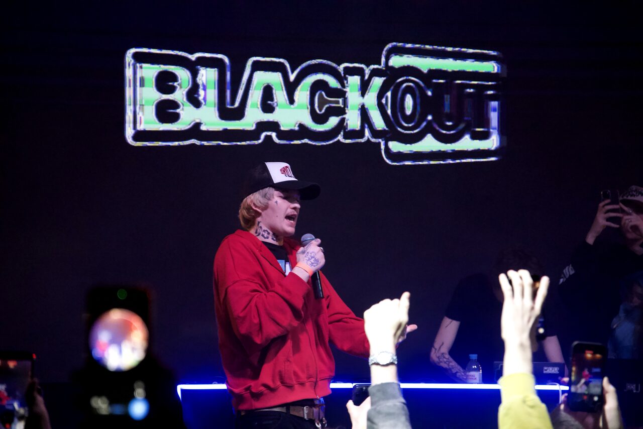 Арендовать Castle Concert Hall(Концертный зал Замок). Концертное пространство с темным интерьером, светящаяся надпись "BLACKOUT", сценическое освещение. На переднем плане человек с микрофоном, окруженный людьми. Используются черные и неоновые цвета.