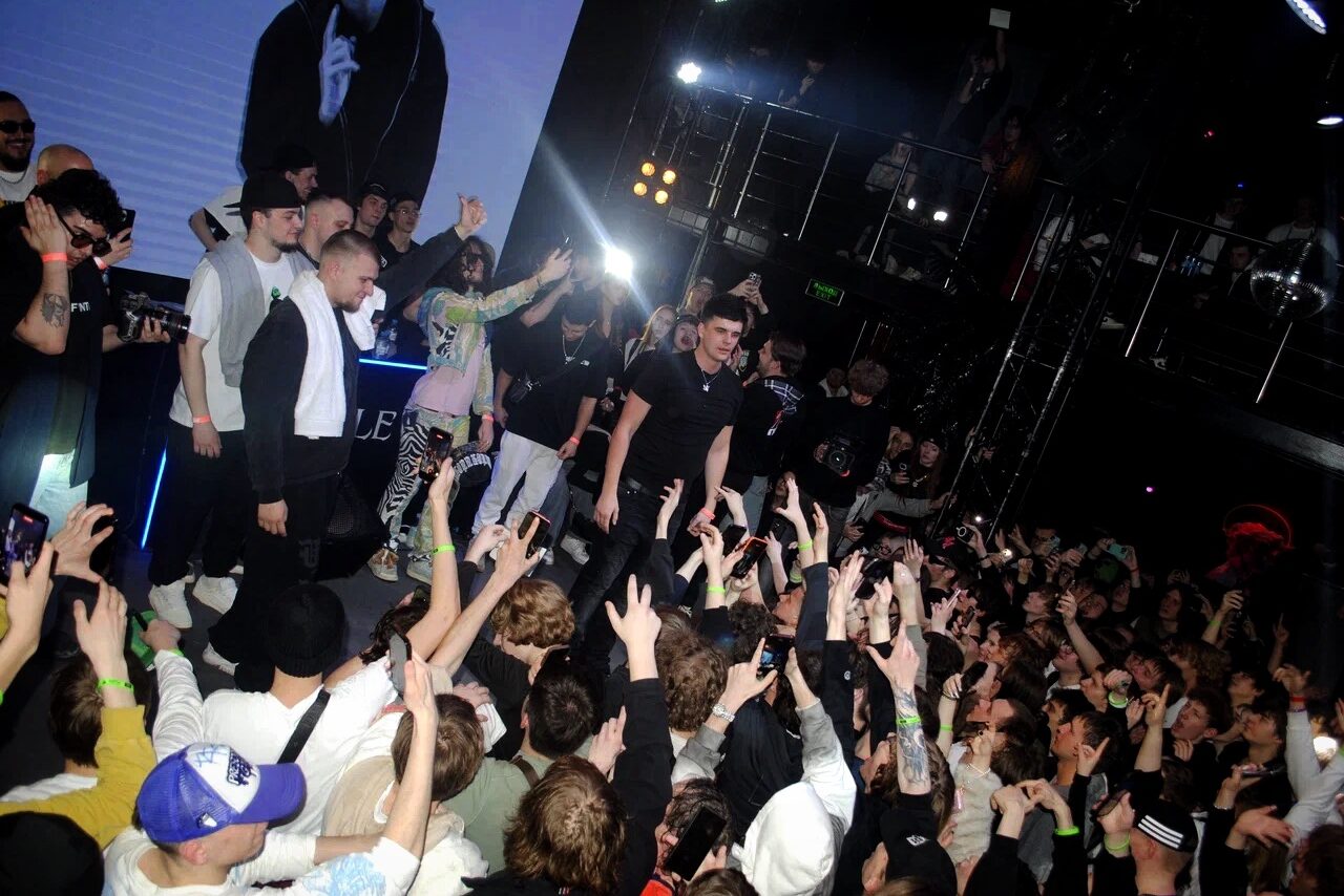 Арендовать Castle Concert Hall(Концертный зал Замок). Клубное пространство с выступающими людьми на сцене перед большой группой аудитории. Многоуровневая планировка с металлическими перилами. Яркое сценическое освещение, темные стены. Участники сцены одеты в повседневную одежду.