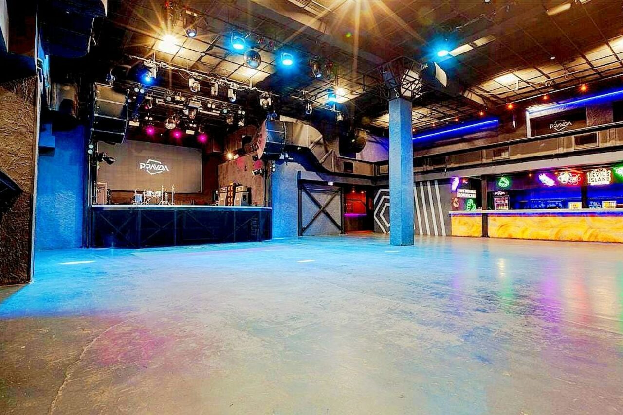 Арендовать PRAVDA(Правда). Просторный зал на 800 гостей. Большое открытое пространство с бетонным полом, сцена с музыкальным оборудованием, барная стойка с подсветкой, разноцветные прожекторы на потолке, металлические конструкции. Потолок с обширной системой освещения и проводки. На экране надпись "PRIZMA".
