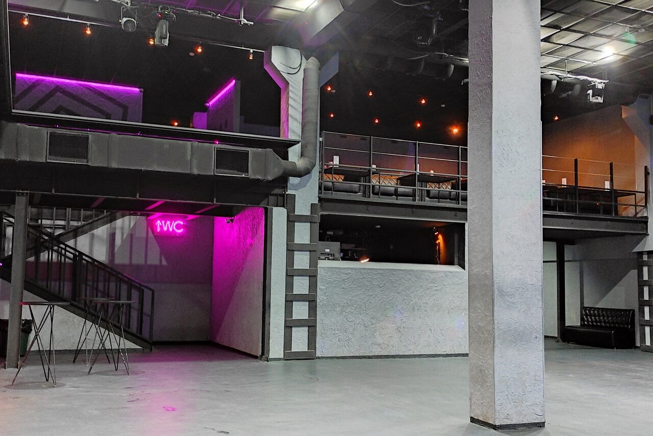 Арендовать PRAVDA(Правда). Просторный зал на 800 гостей. Многоуровневое пространство с бетонным полом, колоннами и балконом. Черная лестница ведет на второй этаж. Неоновая вывеска «WC». Тёмное освещение с точечными светильниками. Стены серые с текстурой. Мебель включает высокие стулья и диваны.