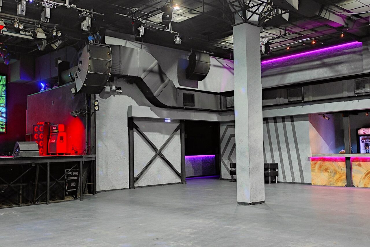 Арендовать PRAVDA(Правда). Просторный зал на 800 гостей. Клубное пространство с бетонным полом и металлическими колоннами. Справа барная стойка с декоративной подсветкой фиолетового и красного цветов. Слева сцена с аудиосистемой. Потолок с подвесными светильниками и колонками. Стены серые с черными элементами.