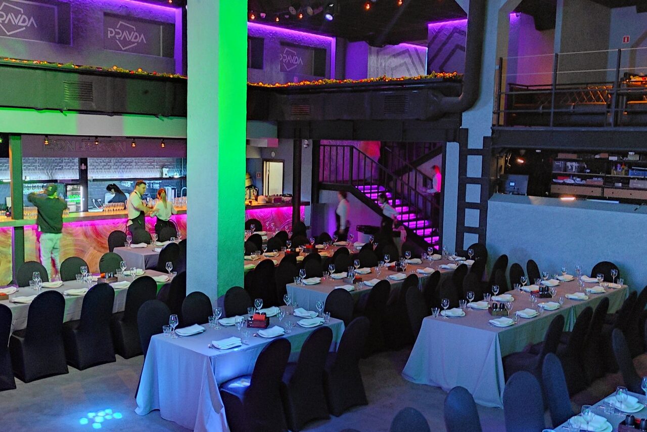 Арендовать PRAVDA(Правда). Просторный зал на 800 гостей. Просторное помещение с двумя уровнями, прямоугольные столы с белыми скатертями, стулья с тёмными чехлами, яркое разноцветное освещение, неоновые элементы, металлическая лестница, знаки с надписью "PANDA" на верхнем уровне, барная стойка с подсветкой.