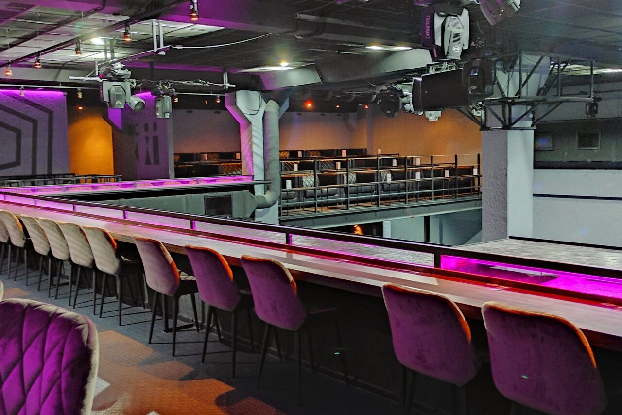 Арендовать PRAVDA(Правда). Просторный зал на 800 гостей. Большое пространство с высоким потолком, длинный барный стол с фиолетовыми барными стульями, разноуровневые платформы. На потолке световые приборы; освещение красного и фиолетового оттенков. Металлические конструкции, белые колонны, черные перила.