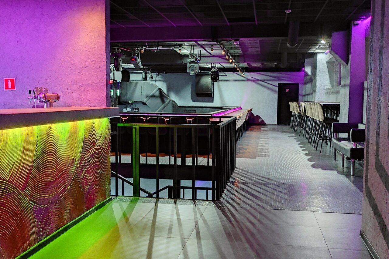 Арендовать PRAVDA(Правда). Просторный зал на 800 гостей. Клубное пространство с барной стойкой, яркой подсветкой фиолетового и зелёного цветов. Черный потолок с вентиляцией и светильниками. Ряды черных стульев вдоль стен. Потолок и стены серые. Пол с серой плиткой. Панель с декоративными узорами.