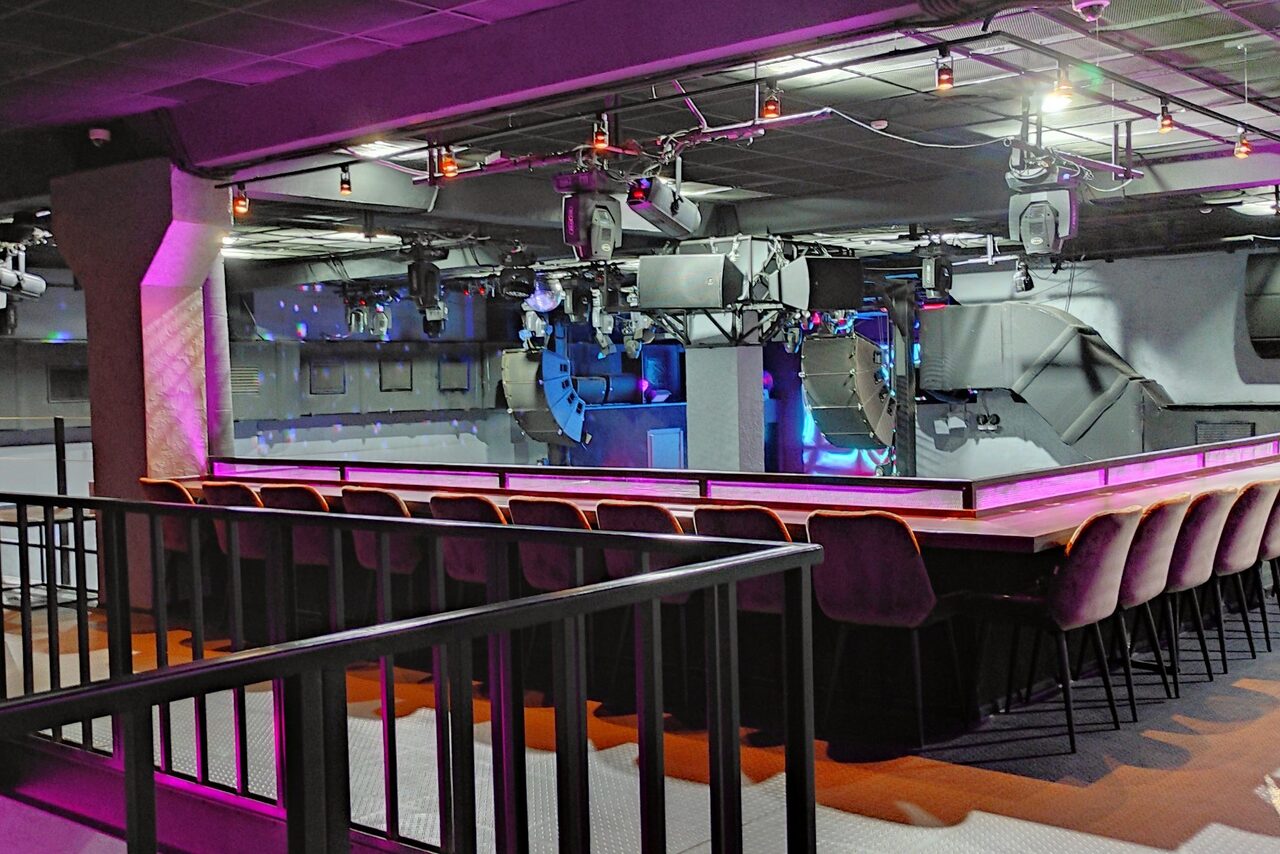 Арендовать PRAVDA(Правда). Просторный зал на 800 гостей. Просторное помещение с черным потолком и освещением. Передний план: барная стойка с мягкими стульями и барными стульями на длинной ножке. В дальнем плане: рулонные динамики и световое оборудование. Стены серые, с металлическими элементами.