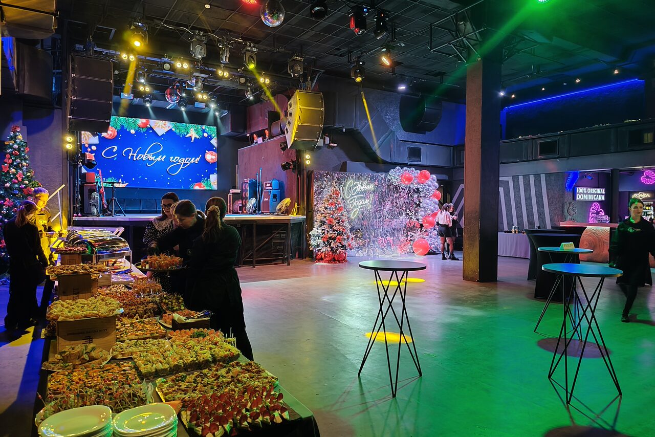 Арендовать PRAVDA(Правда). Просторный зал на 800 гостей. Большое помещение с высокой сценой, оборудованной аудиосистемой и экранами со световыми эффектами. В центре зала высокие круглые столы, слева стол с разнообразной едой. Освещение разноцветное, декорации с новогодней тематикой.
