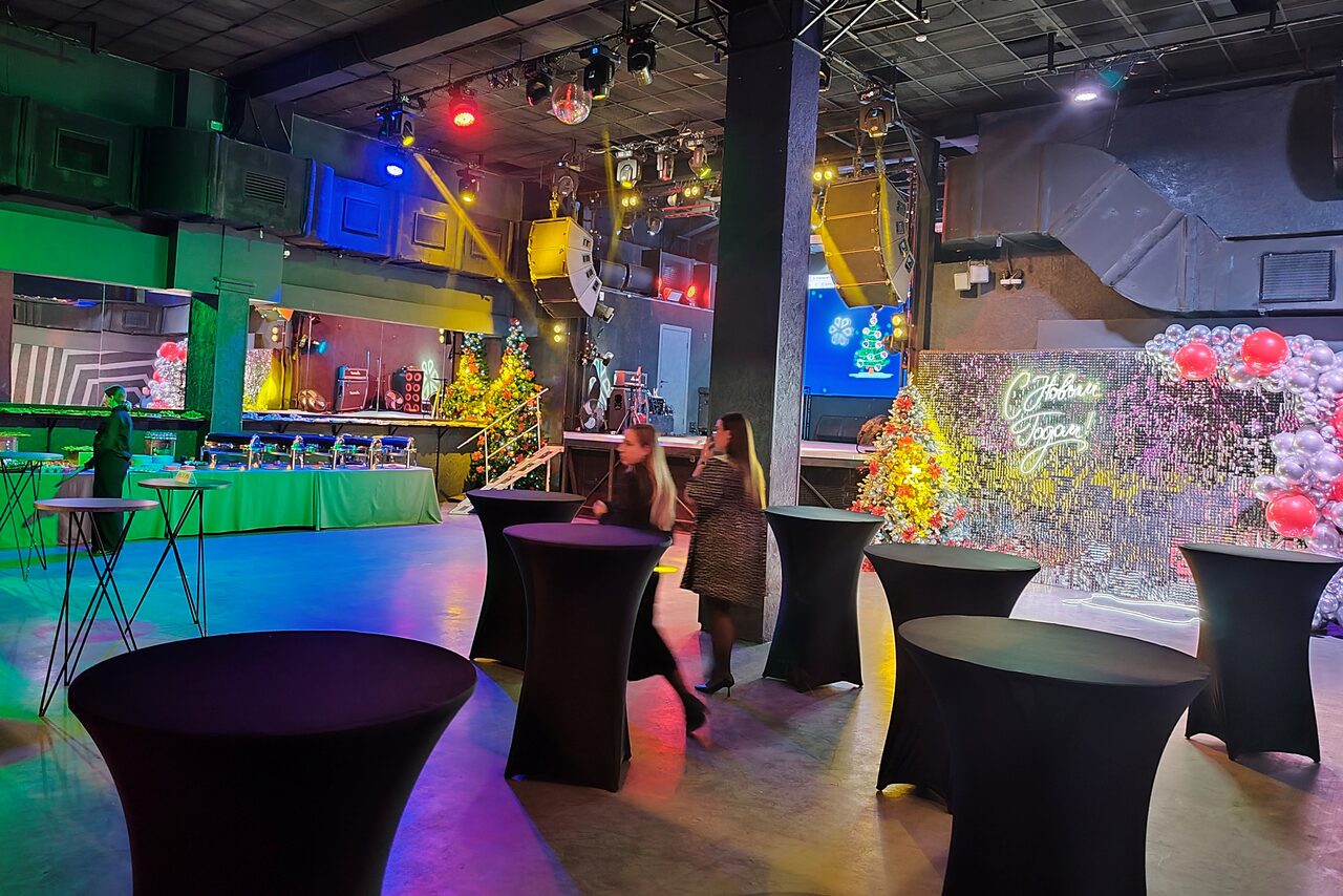 Арендовать PRAVDA(Правда). Просторный зал на 800 гостей. Большое пространство с ярким освещением, включающим цветные прожекторы. На полу расставлены круглые высокие столы черного цвета. На заднем плане стол с зеленой скатертью и декорации с шарами и неоновой надписью. Потолок с индустриальными элементами.