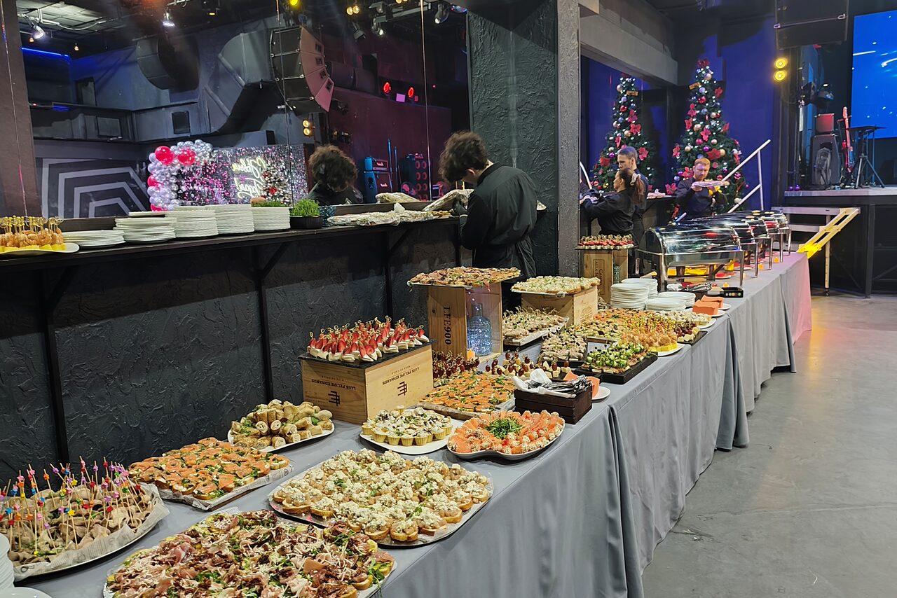 Арендовать PRAVDA(Правда). Просторный зал на 800 гостей. Большое пространство с серыми стенами и потолком, высокий банкетный стол с серой скатертью, уставленный разнообразными блюдами на подносах и деревянных ящиках. На заднем плане новогодние ёлки, сцена, освещение разного цвета.