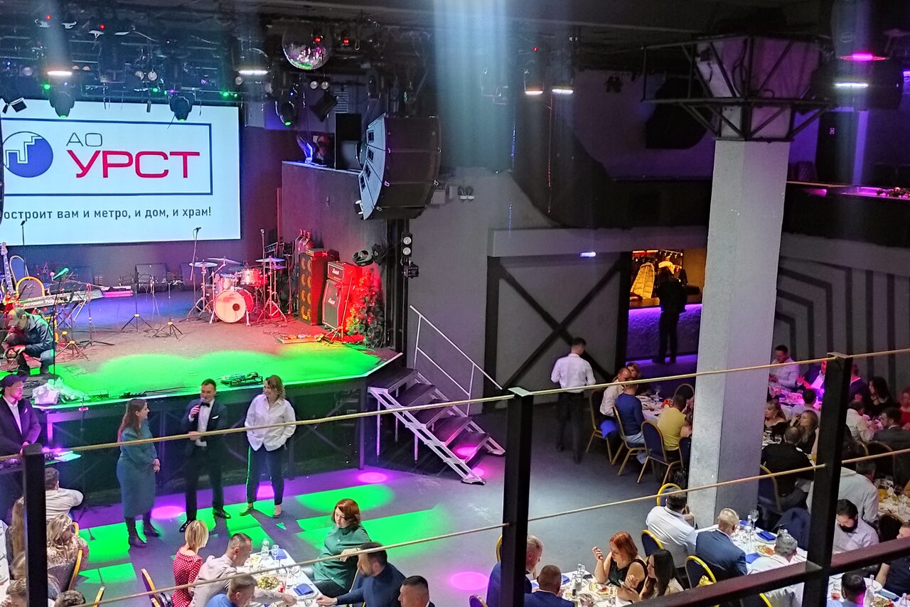 Арендовать PRAVDA(Правда). Просторный зал на 800 гостей. Сцена с музыкальными инструментами и светооборудованием, стулья и столы с людьми, разноцветная подсветка, крупный экран с текстом "АО УРСТ", леера вокруг зоны, серый и фиолетовый интерьер.