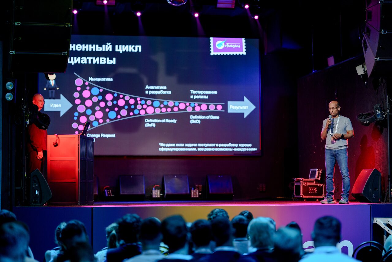 Арендовать PRAVDA(Правда). Просторный зал на 800 гостей. Сцена с двумя выступающими, большой экран с диаграммой и текстом, перед сценой сидят люди. Освещение цветное, голубые и фиолетовые тона. На сцене стоят колонки, сбоку виден ноутбук. Текст на экране и детали диаграммы.