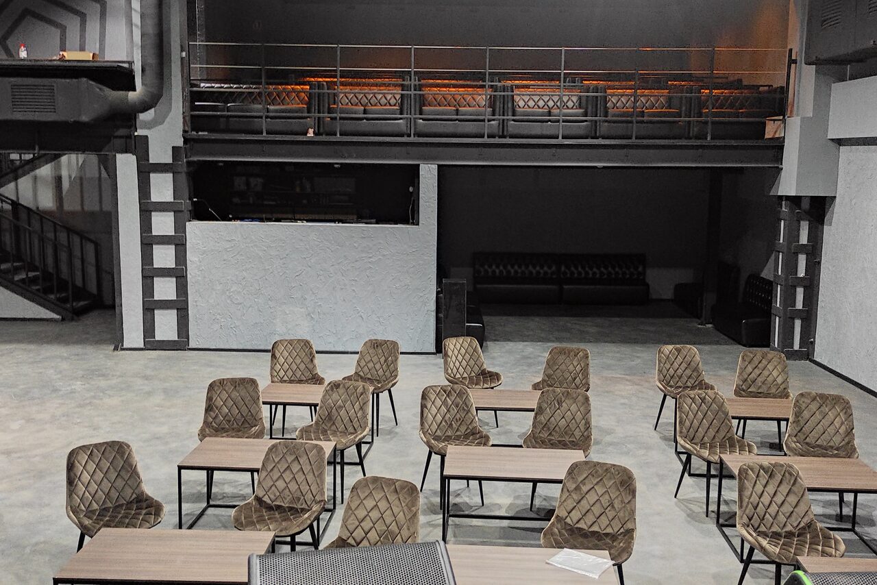 Арендовать PRAVDA(Правда). Просторный зал на 800 гостей. Просторное помещение с бетонным полом. Двенадцать стульев и шесть столов, расположенные в два ряда. Справа лестница на второй этаж с металлическими перилами. Стены серого цвета, сверху металлические балки. Тёмные кожаные диваны на заднем плане.