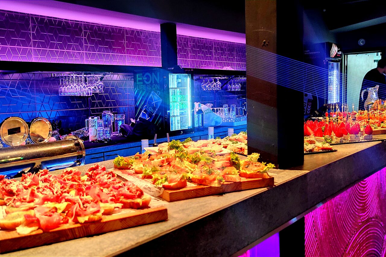 Арендовать PRAVDA(Правда). Просторный зал на 800 гостей. Барное пространство с высокой стойкой, на которой размещены блюда с закусками. На заднем плане барная зона с висящими бокалами, бутылками и подсветкой. Цвета: фиолетовый, черный. Используются неоновые подсветки и геометрический узор на стенах.