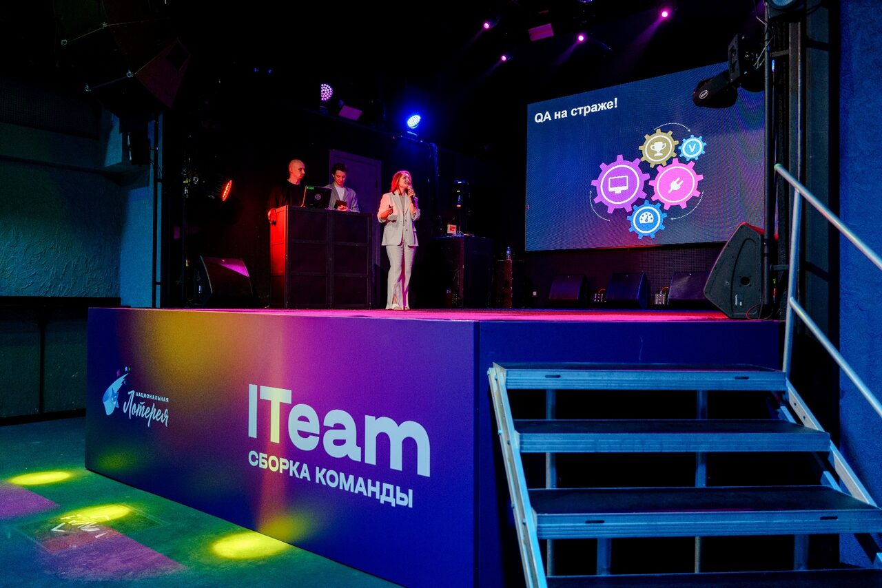 Арендовать PRAVDA(Правда). Просторный зал на 800 гостей. Сцена с двумя людьми и экраном с графикой, черные колонки по бокам, рядом лестница. Фиолетовая подсветка, синий фон сцены. На переднем плане текст «ITeam Сборка команд».
