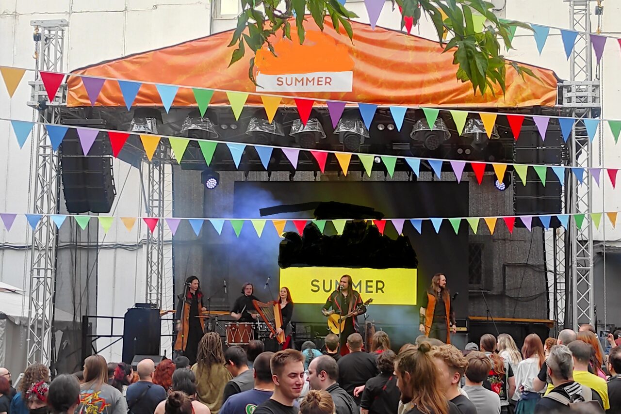 Арендовать PRAVDA(Правда). Просторный зал на 800 гостей. Открытая сцена с оранжевым навесом и надписью "SUMMER". Концертная площадка с пятью музыкантами на сцене, возле которых расположены барабаны и гитара. Над сценой гирлянды разноцветных флажков. Большая толпа людей перед сценой.