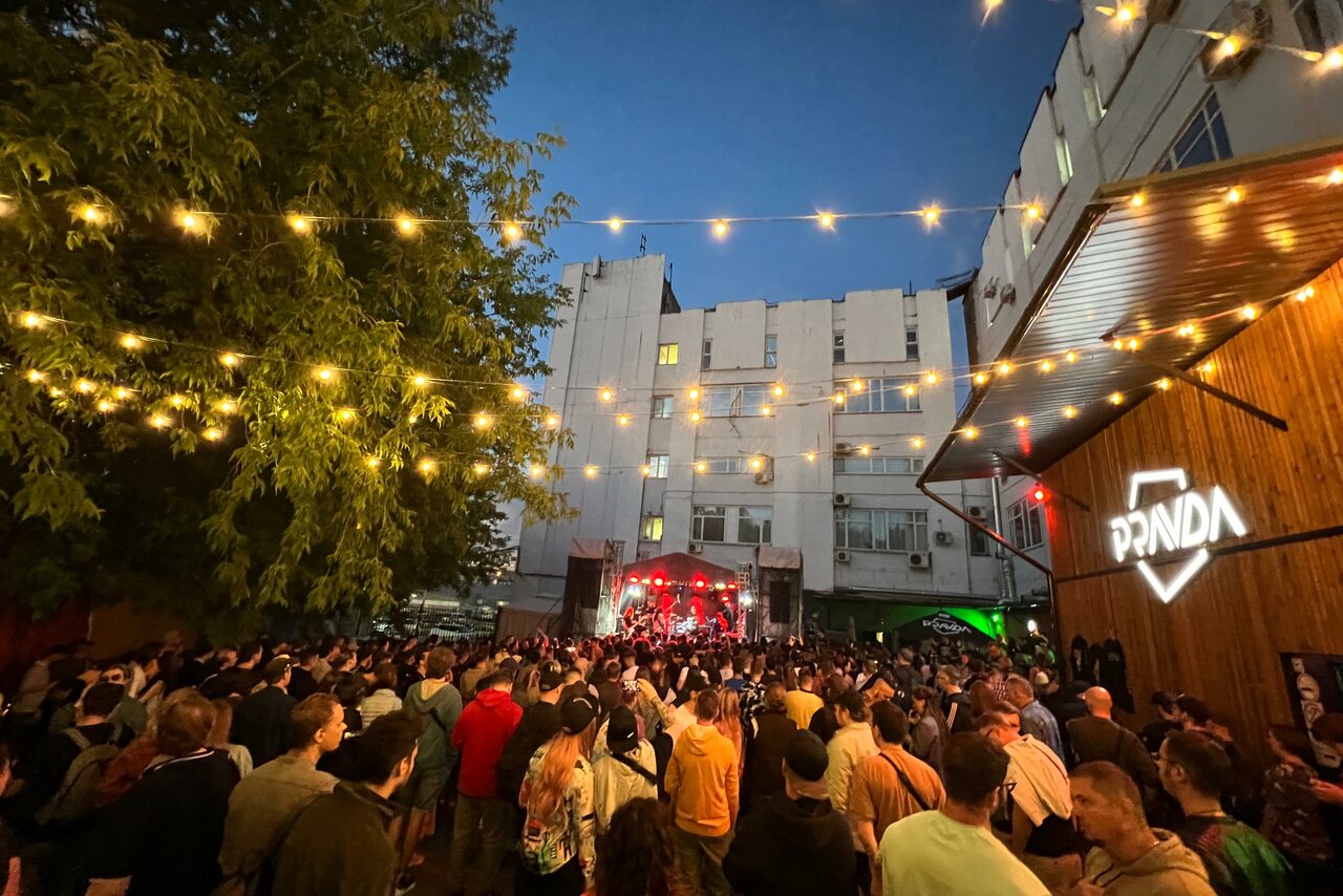 Арендовать PRAVDA(Правда). Просторный зал на 800 гостей. Открытая площадка с толпой людей, сцена с музыкантами, фонарики над площадкой, белое здание на заднем плане, зелень, вывеска с текстом "PRANDA" на деревянной стене, вечернее время.