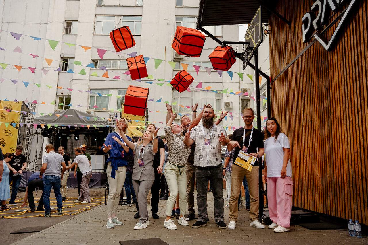 Арендовать PRAVDA(Правда). Просторный зал на 800 гостей. Открытое пространство во дворе с бетонным покрытием, задний фон с многоэтажным зданием и праздничными гирляндами. Многоцветные флажки и кубы с рисунком баскетбольных мячей. Группа людей перед деревянной стеной, портативная сцена с музыкальным оборудованием.