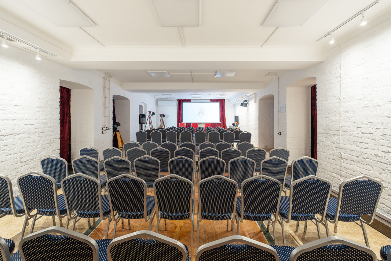 Арендовать Светлый Лофт. Пространство для мероприятий с белыми кирпичными стенами и деревянным полом. Ряды черных стульев с серыми вставками, направленных к белому экрану проектора. Потолок с точечным освещением, в задней части помещения установлены штативы для камер.