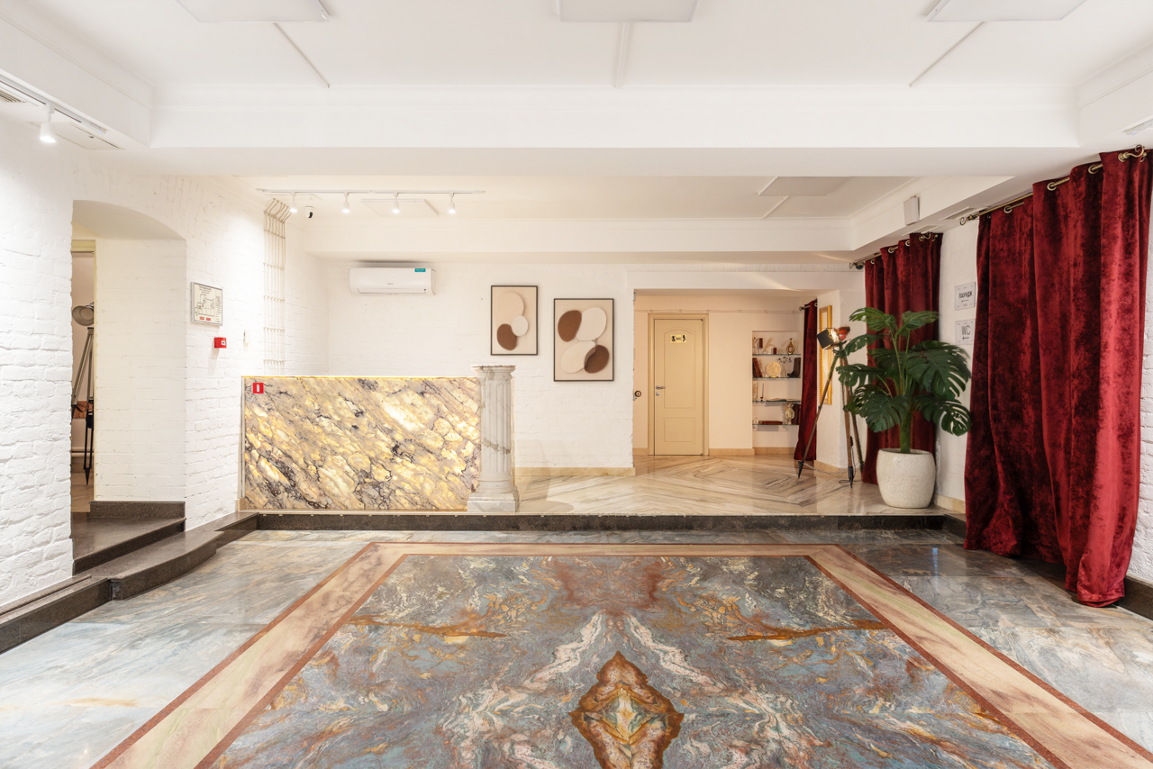 Арендовать Светлый Лофт. Просторное помещение с мраморным полом и стойкой из камня. Белые кирпичные стены, красные шторы, картины на стене. Растение в горшке. Освещение с помощью встраиваемых светильников. Кондиционер на стене справа.