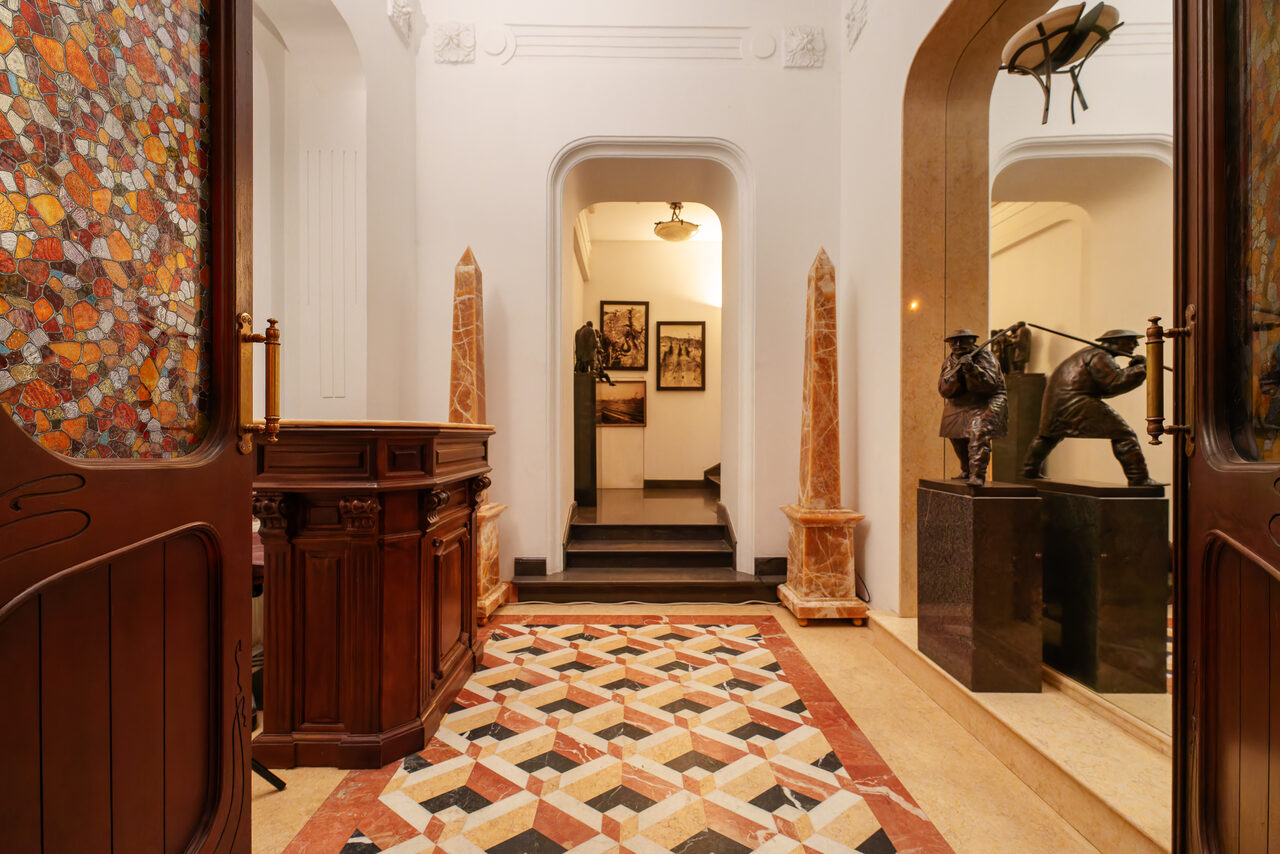 Арендовать Светлый Лофт. Коридор в интерьере с мозаичным полом в геометрическом узоре, деревянной панелью с витражами, скульптурами на постаментах, мраморными украшениями. Белые стены, арочные проемы, декоративное освещение, фотографии на задней стене.