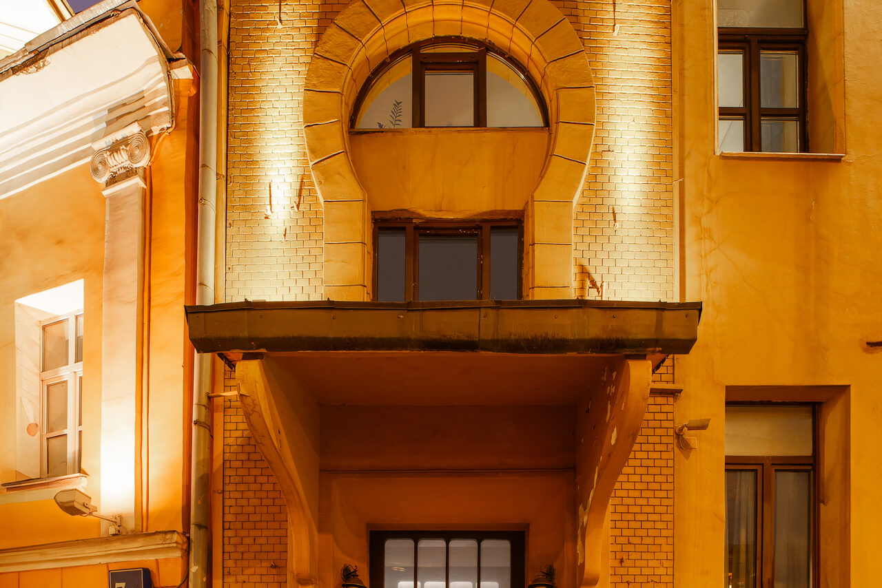 Арендовать Светлый Лофт. Фасад здания с аркой над тёмной входной дверью. Стены отделаны светлой кирпичной кладкой и штукатуркой. Фасадное освещение подчёркивает архитектурные элементы. Окна сверху и слева от входа. Вечернее освещение.