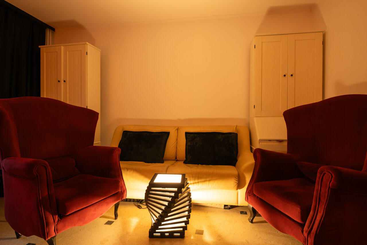 Арендовать Светлый Лофт. Комната с мягким освещением, диван, два кресла бордового цвета, между ними светильник на столе, два шкафа с дверцами, светлые стены, темные подушки на диване.