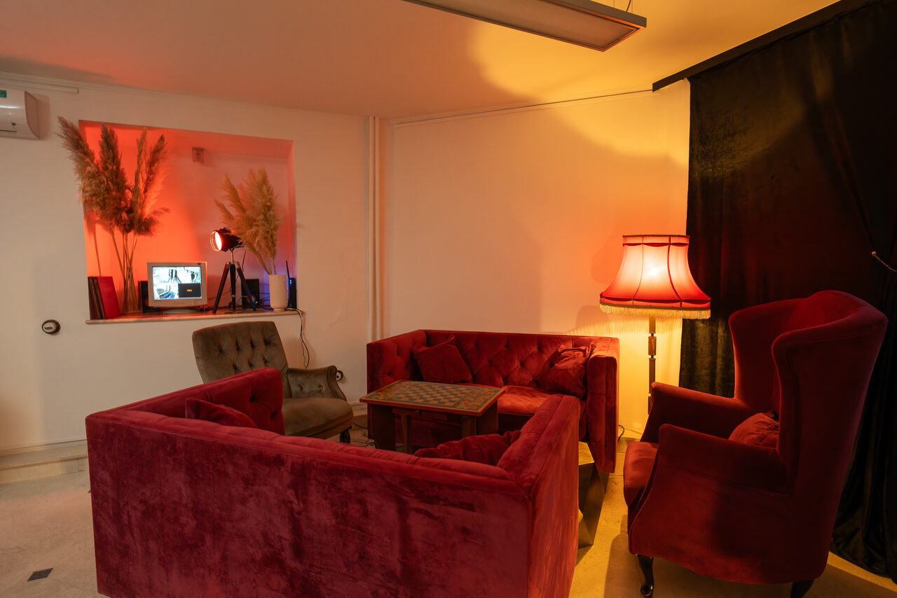 Арендовать Светлый Лофт. Просторная комната с мягкой мебелью: два красных дивана и кресло, деревянный столик. Освещение от настольной лампы с абажуром. На заднем плане компьютер и декоративные растения. Стены белые, пол светлый. На окне черная занавеска.