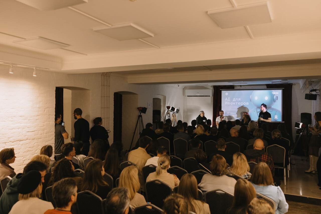 Арендовать Светлый Лофт. Аудитория с людьми, расположенными на стульях рядами. На сцене выступающие за столом. Белые кирпичные стены, светло-бежевая окраска потолка с прямоугольными светильниками. На экране текст "для" с изображением камеры. Камеры на штативах у сцены.