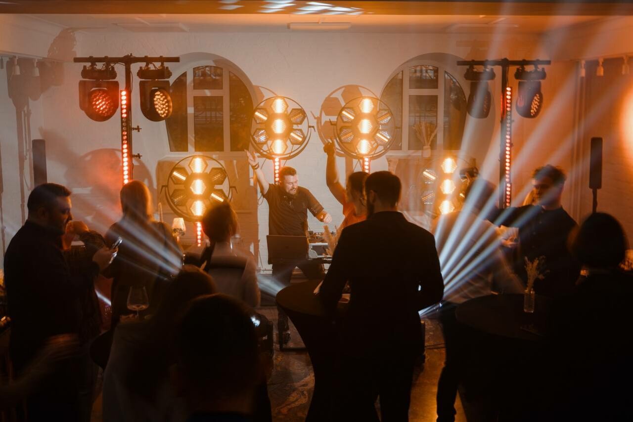 Арендовать Светлый Лофт. Танцевальная площадка с мягким освещением, круглыми столами, цветочными вазами, сценой и диджейским оборудованием. Световые прожекторы, лучи света. Интерьер в тёмных и светлых тонах, окна на заднем плане.