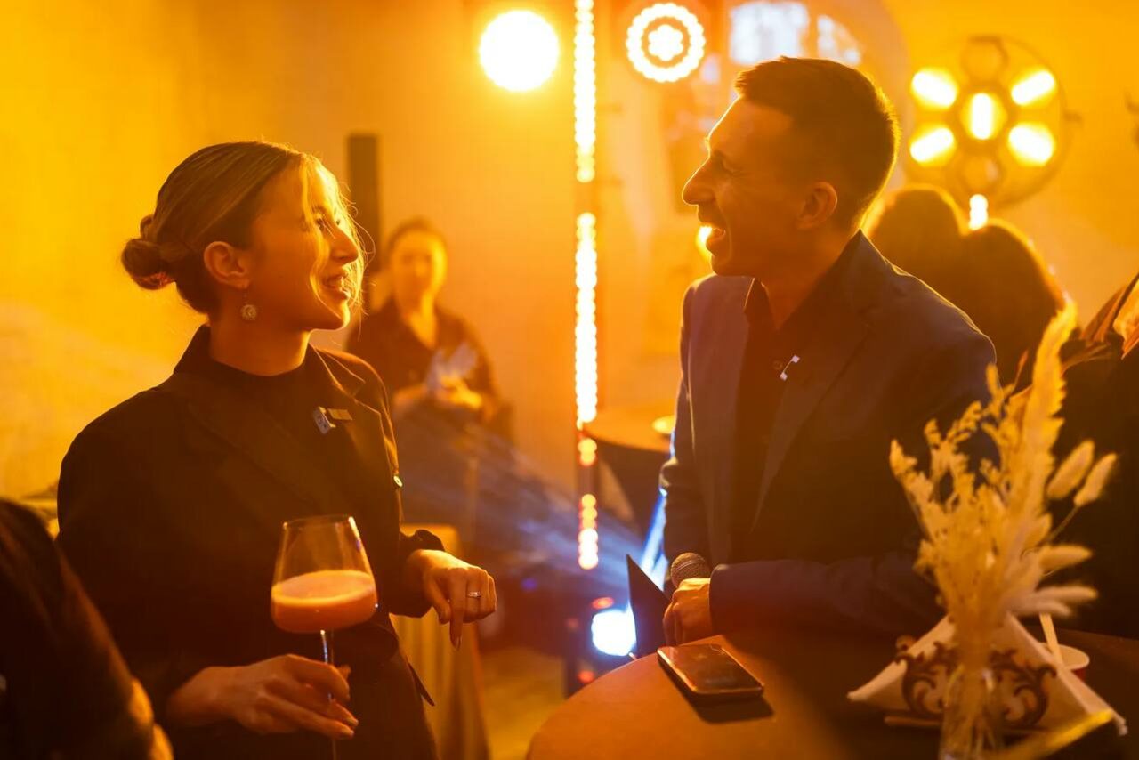 Арендовать Светлый Лофт. Пространство с теплым освещением, несколько человек стоят рядом с барной стойкой. На столе бокал с напитком и небольшой декор из сухоцветов. На заднем плане видны световые приборы. Элементы интерьера из дерева и металла.