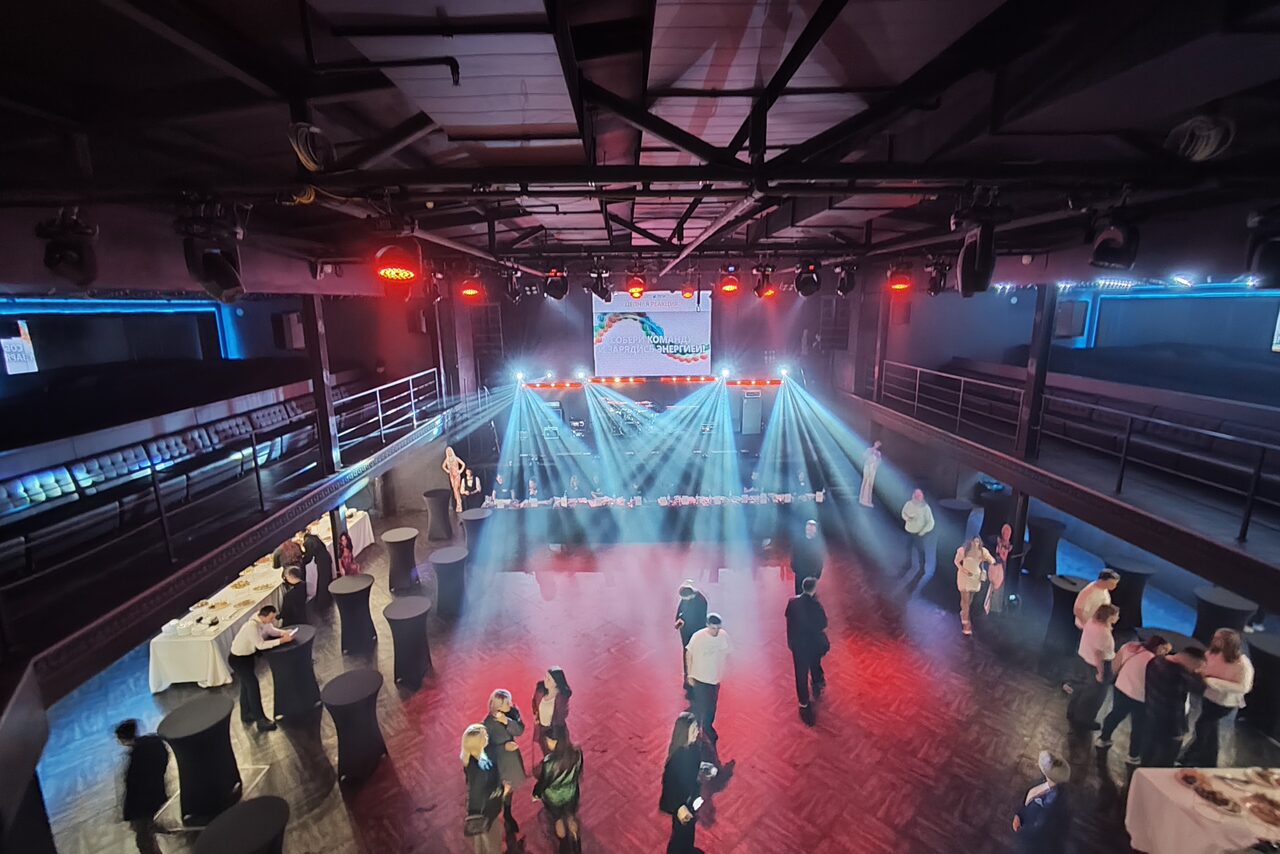 Арендовать СВОБОДА. Зал на 1200 человек. Просторная комната с высокими потолками, оснащена сценой с подсветкой и большим экраном. Вокруг расставлены высокие круглые столы с белыми скатертями. На полу паркет, по периметру полумрак с ярким светом на сцене.