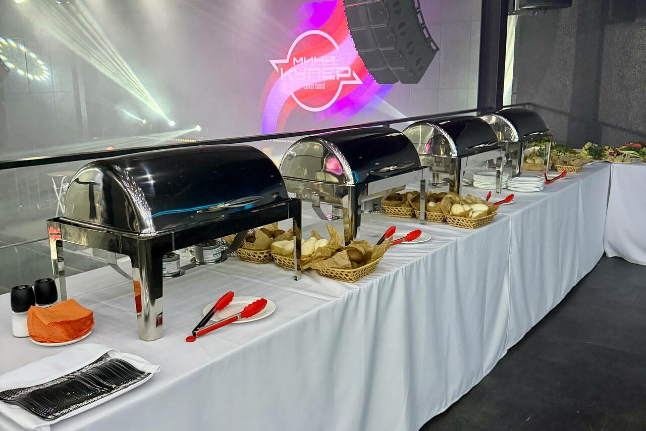 Арендовать СВОБОДА. Зал на 1200 человек. Помещение для банкета с длинным столом, покрытым белой скатертью. На столе четыре металлических контейнера для еды, подносы с хлебом, красные щипцы, посуда, посолонки и салфетки. На заднем плане светодиодные панели с текстом.