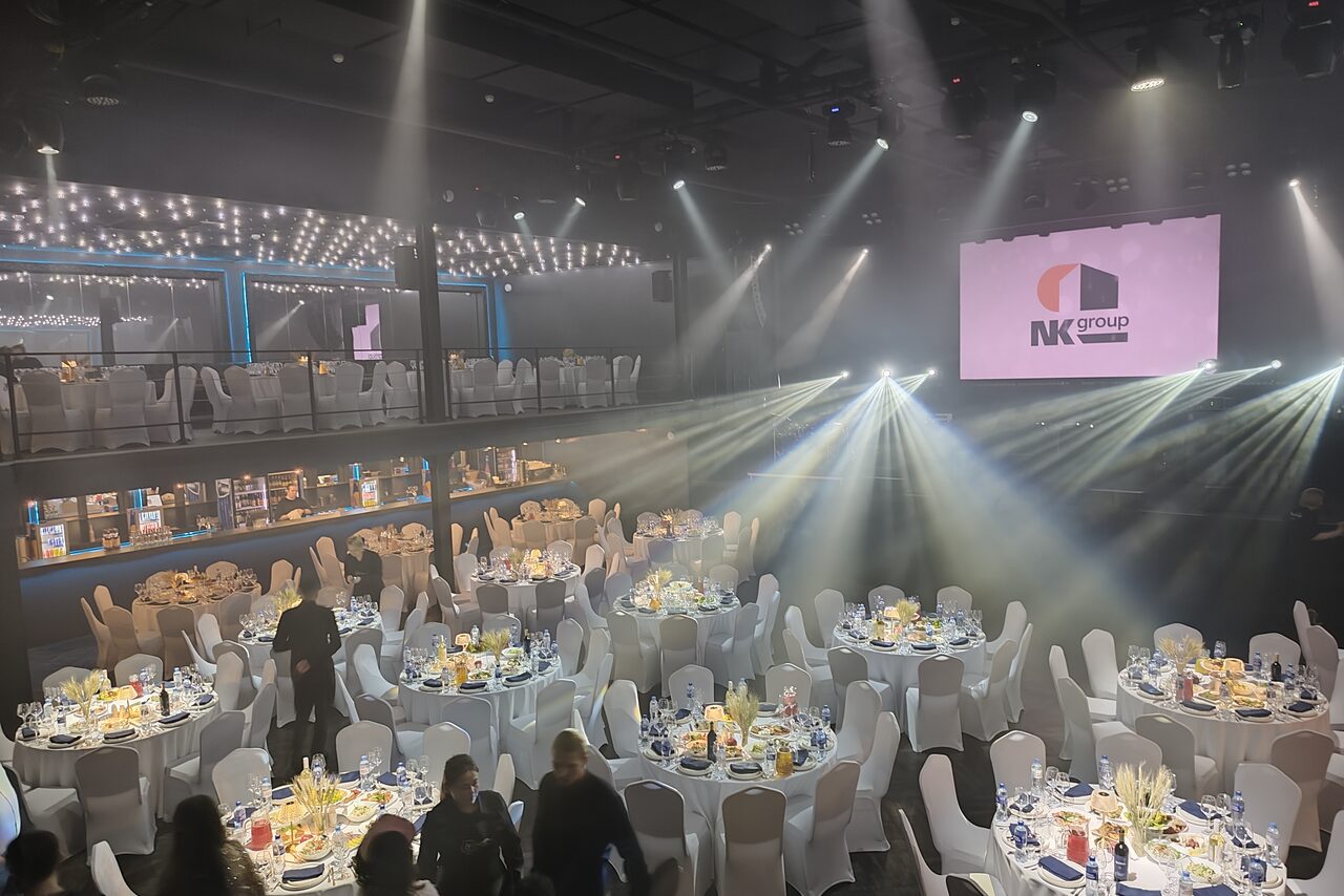 Арендовать СВОБОДА. Зал на 1200 человек. Большой зал с круглым столами, покрытыми скатертями; на каждом столе свечи и сервировка. Белые стулья, световые лучи по залу. Двухуровневое пространство, на верхнем уровне также столы и стулья. На стене висит экран с логотипом.