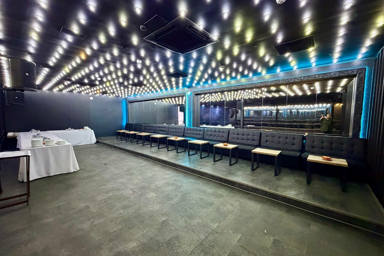 Арендовать СВОБОДА. Зал на 1200 человек. Помещение с затемненным интерьером, черные стены и потолок с множеством встроенных светильников. Справа вдоль стены длинные серые скамьи с подушками и маленькими столиками. Слева накрыт белый стол. Пол покрыт темной плиткой.