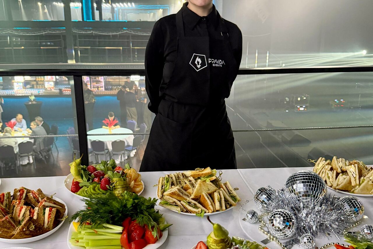 Арендовать СВОБОДА. Зал на 1200 человек. Человек в черной униформе стоит за столом с разнообразными закусками и фруктами. На столе также лежат зеркальные диско-шары. На заднем плане видна часть банкетного зала с накрытыми столами и гостями. Освещение яркое.