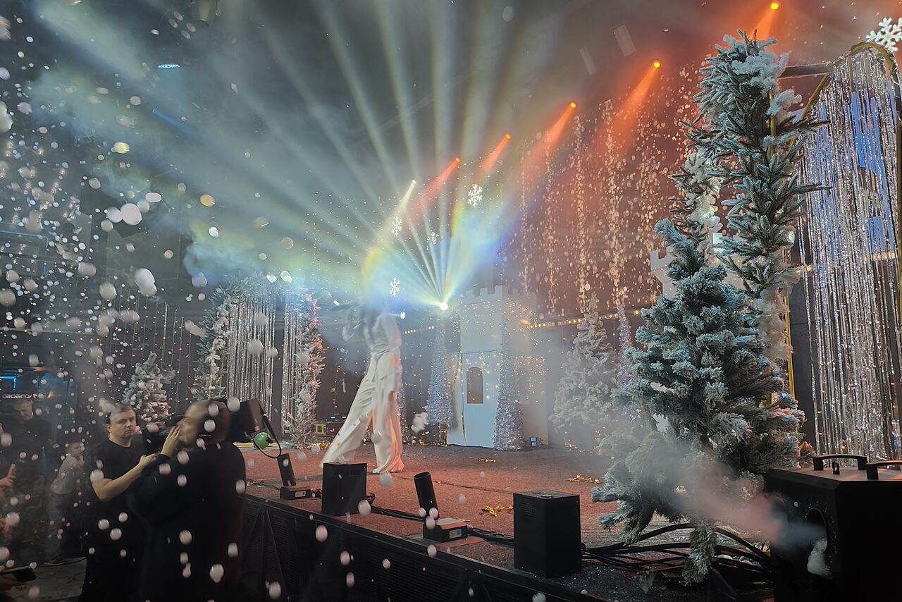 Арендовать СВОБОДА. Зал на 1200 человек. Сцена с новогодним декором, украшенная искусственными елками и снежинками. Световое шоу с разноцветными прожекторами и мигающими гирляндами. Перед сценой микрофоны и пульт для управления светом. Деревья покрыты "снегом".