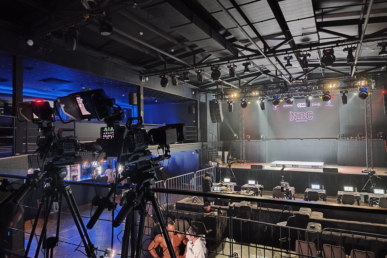 Арендовать СВОБОДА. Зал на 1200 человек. Концертный зал с камерой на штативе, сцена с фоновой панелью и надписью "NBC". Верхнее освещение с софитами, несколько светодиодных экранов. Пространство многоуровневое, сиденья расставлены на арене и балконе. Тёмная цветовая гамма.