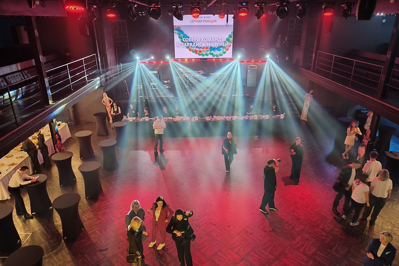 Арендовать СВОБОДА. Зал на 1200 человек. Большое внутреннее пространство с деревянным полом и балконами на втором уровне. На переднем плане высокие круглые столы. На заднем плане сцена с экраном и световыми эффектами. Люди стоят и общаются. Освещение разноцветное.