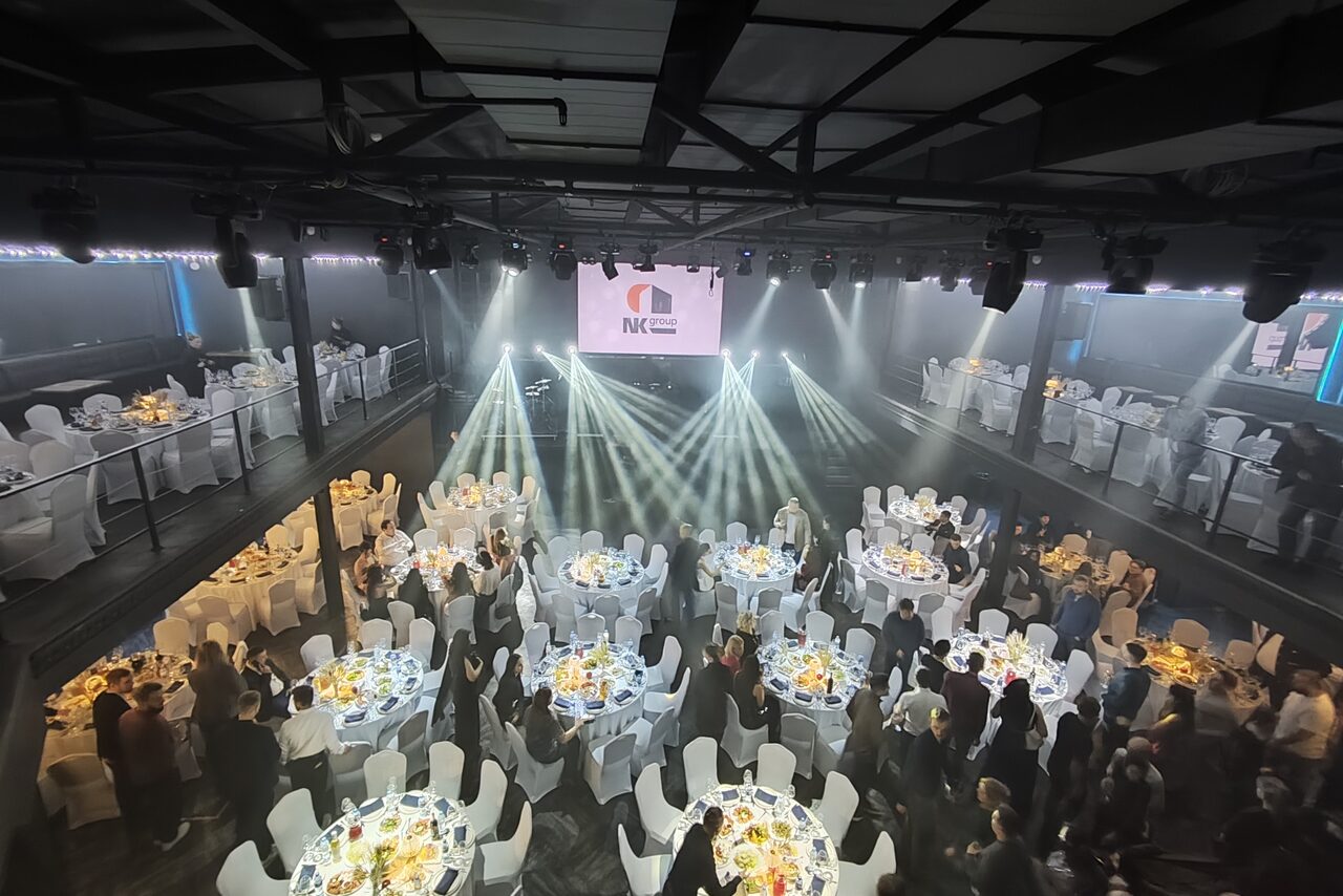 Арендовать СВОБОДА. Зал на 1200 человек. Большое мероприятие в закрытом пространстве, несколько круглых столов с белыми скатертями и стульями, блюда с едой на столах, люди в вечерней одежде. Сцена с экраном и световыми прожекторами. Верхний ярус с дополнительными местами.