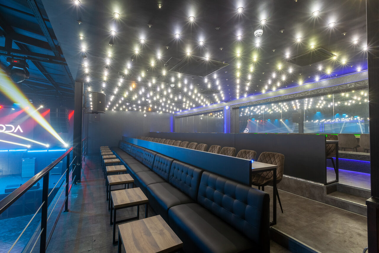 Арендовать СВОБОДА. Зал на 1200 человек. Современное пространство с амфитеатром и рядами темных диванов и деревянных столиков. Потолок украшен множеством светодиодных ламп. Пол бетонный, интерьеры выполнены в серых и черных тонах с элементами цветной подсветки.