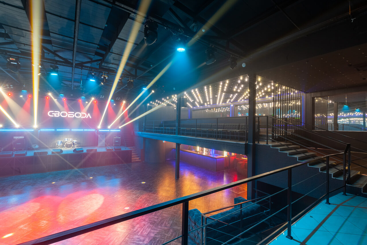 Арендовать СВОБОДА. Зал на 1200 человек. Пространство для мероприятий с деревянным полом и сценой, освещение разноцветными прожекторами, подвесные светильники на потолке, металлические перила, лестница, надпись на заднем плане, открытая планировка с балконом.