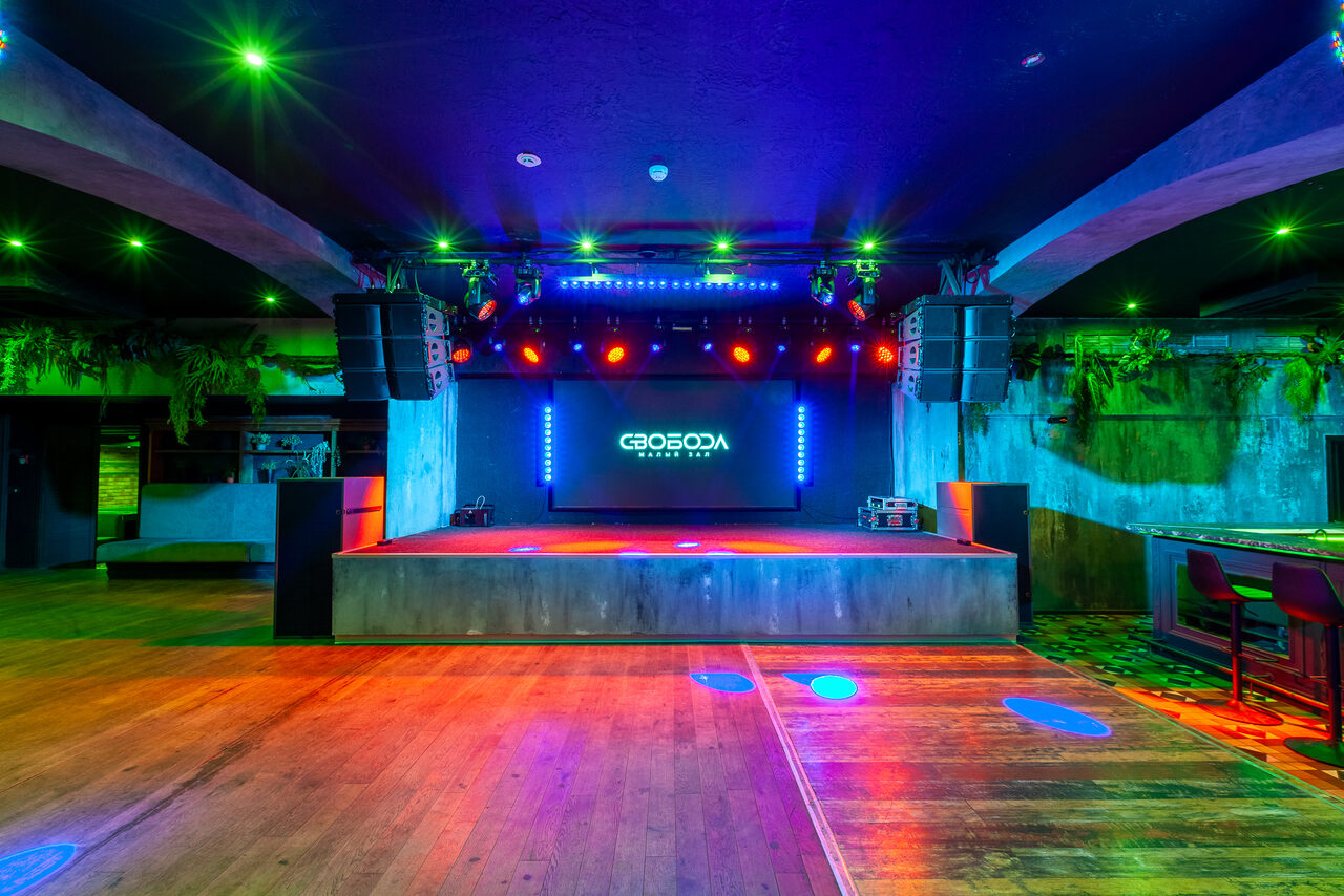 Арендовать СВОБОДА. Зал на 1200 человек. Большое помещение с деревянным полом и сценой, оборудованное звуковыми системами и освещением различных цветов. Наверху сцены светодиодный экран с белым текстом. Справа барная стойка с высокими стульями. Стены текстурные, темные.